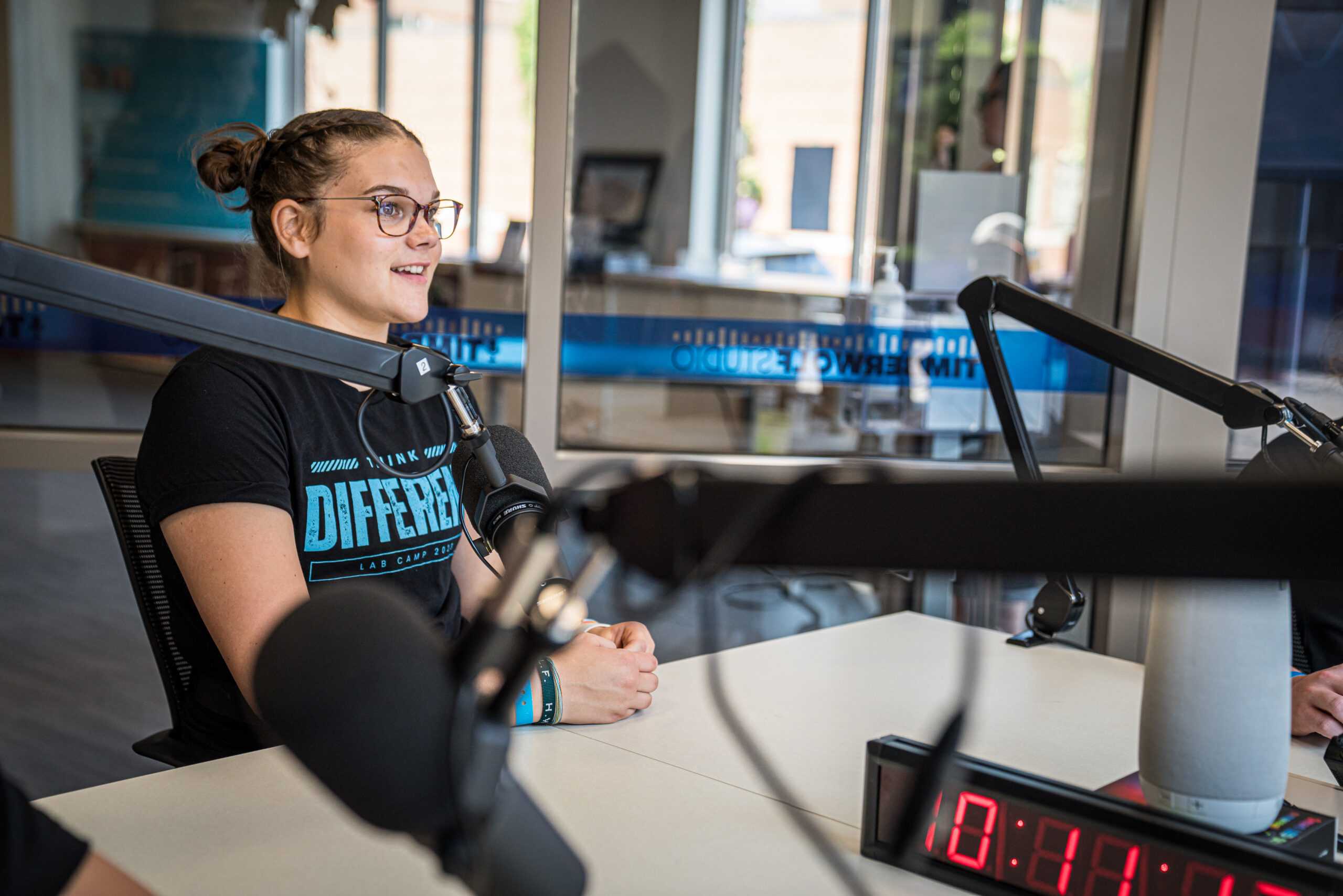 A young woman in glasses and wearing a bun sitting in the podcast studio, talking into a microphone.