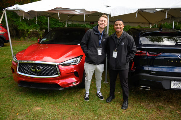 Two men, embodying the spirit of undergraduate academics, stand smiling between a red and a black car under a canopy at an outdoor event.