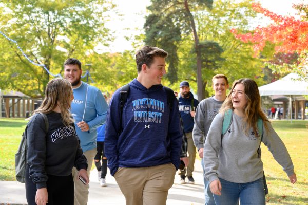 A group of young adults, possibly discussing undergraduate scholarships and grants, strolls together on a sunny day. They are clad in casual sweatshirts with backpacks slung over their shoulders, enveloped by trees and a lush grassy area.