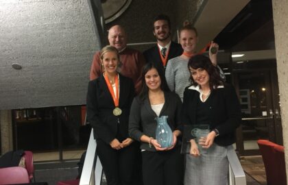 A group of six people in business attire stand on stairs at Northwood University, smiling after their big wins. Some are holding awards and wearing medals around their necks. A modern building interior is visible in the background, reflecting their success in competitive speech.