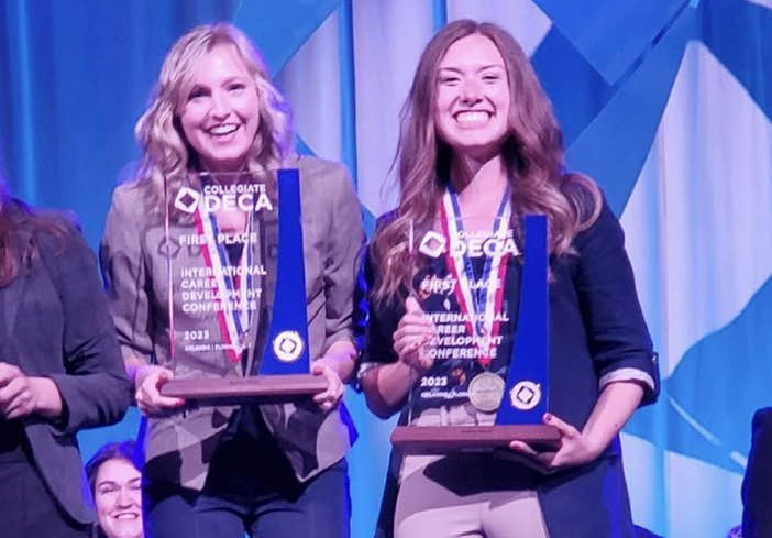 Two individuals are smiling and holding awards on a stage at an international DECA conference.