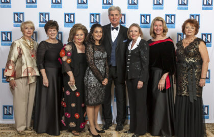 A group of eight people dressed in formal attire stands in front of a backdrop with a repeating logo pattern at a formal event.