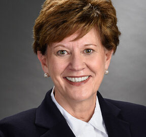 A woman with short brown hair, smiling, wearing a dark blazer, a white shirt, and pearl earrings, against a gray background.