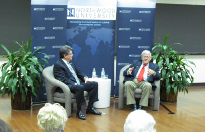 Two men are seated on a stage during Northwood University's Values Emphasis Week, with a banner and potted plants in the background. One man gestures as he speaks, delving into Northwood's history, while the other listens attentively.