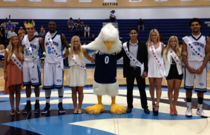 A group of six people and a mascot pose on a basketball court. Two men and one woman wear sashes and are crowned. The others wear sashes but no crowns. Bleachers are in the background.