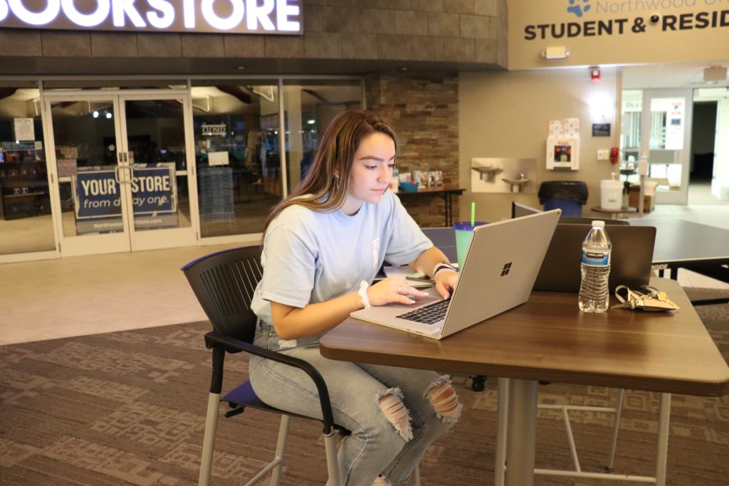A person in a blue shirt focuses on their laptop at a student center table, balancing academics with ease. Nearby, a water bottle and cup provide refreshment during this dedicated study session.