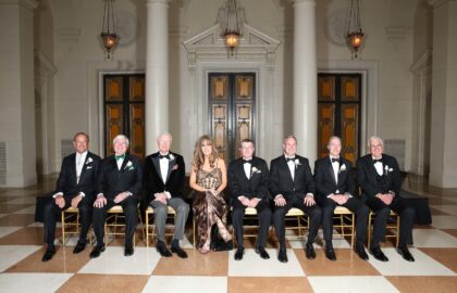 Seven men and one woman are seated in a row on chairs in a formal setting with ornate doors in the background. The men are wearing tuxedos, and the woman is in a patterned dress.