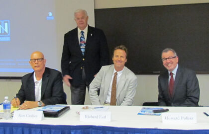 Four men are at a table for a panel discussion. Three of them sit while one stands behind them. Name placards and materials are on the table. A projection screen and blackboard are in the background.