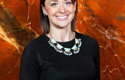 A woman with shoulder-length brown hair, wearing a black top and a statement necklace, stands in front of an orange and black marbled background, smiling at the camera.