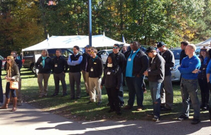 A group of people stands outdoors on a sunny day at Northwood University's International Auto Show, listening attentively. Tents and trees provide a picturesque backdrop.