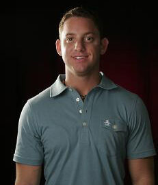 A person wearing a blue polo shirt stands against a dark background, facing the camera and smiling.