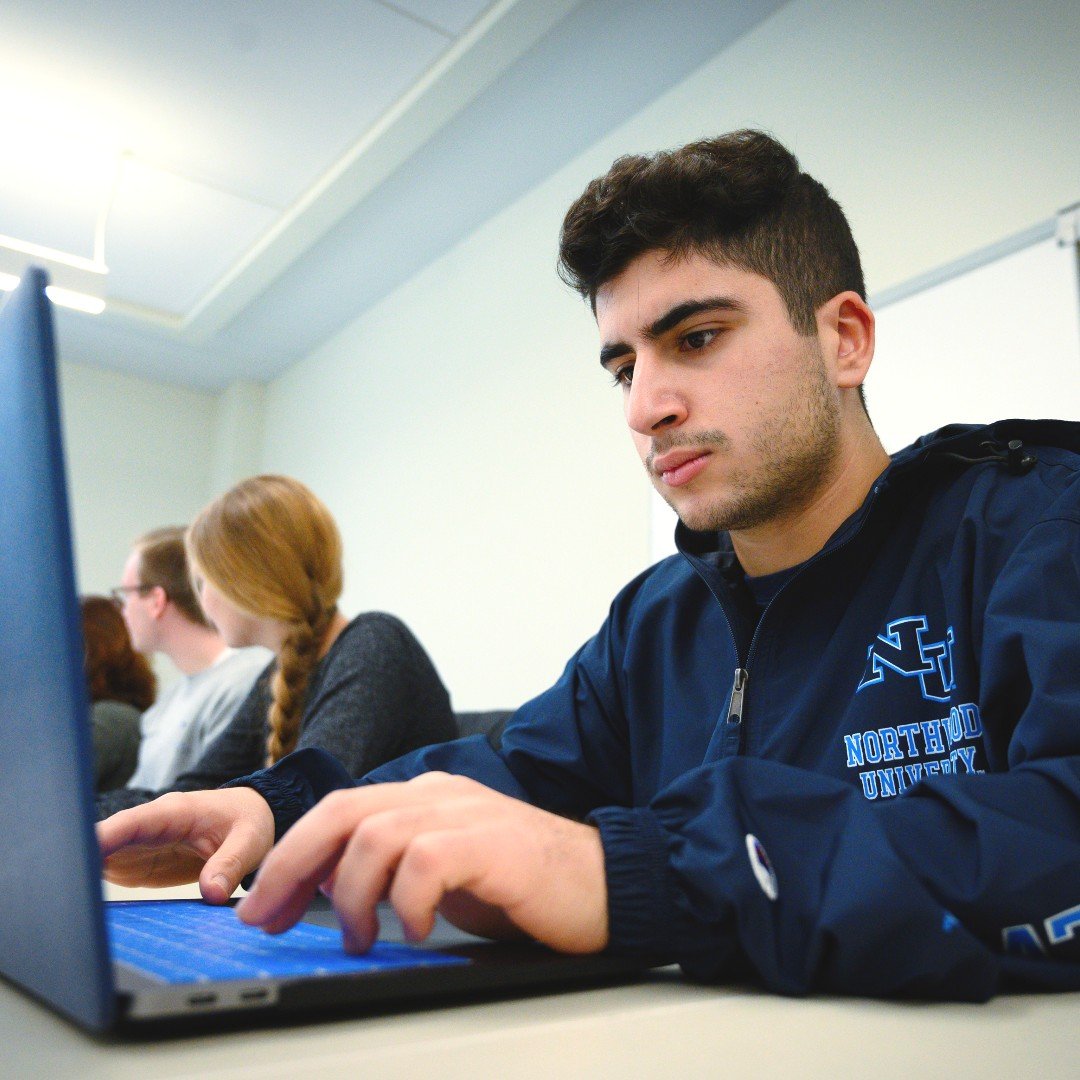 A person in a Northwood University jacket launches into work on a laptop in a classroom setting, while others are seated in the background.