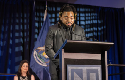 A man with dreadlocks in a suit speaks at a podium, with a woman seated behind him on stage and a flag in the background.