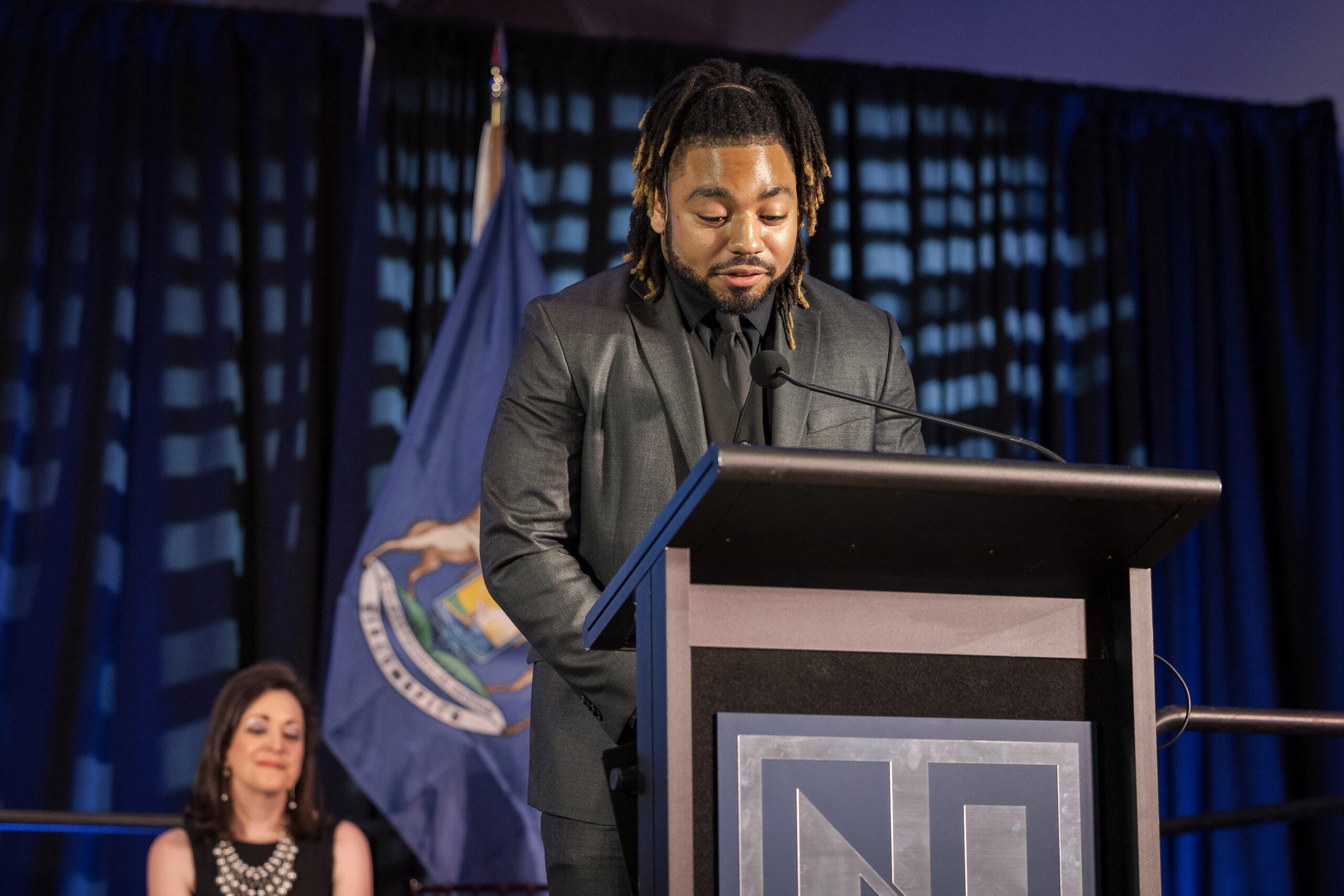 A man with dreadlocks in a suit speaks at a podium, with a woman seated behind him on stage and a flag in the background.