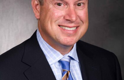 A man with short gray hair wearing a suit and blue striped tie is smiling against a dark gray backdrop.