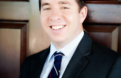 A person wearing a dark suit, white shirt, and striped tie is smiling in front of a wooden door.