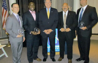 Five men in formal attire stand next to each other, holding awards in recognition of their achievements. An American flag and a banner are visible in the background.