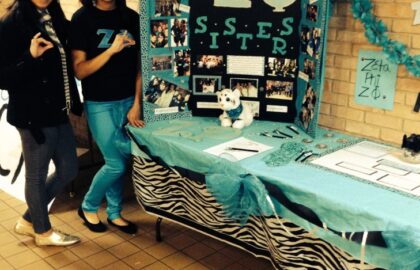 Two people stand beside a decorated booth displaying photos and information about a sorority, with "Zeta Phi" and "Sisters" prominently featured on the board.