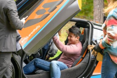 A person is smiling while seated in a sports car with its door open. Several people stand nearby. The car has an orange and blue design with the hashtag #McLarenRally visible.