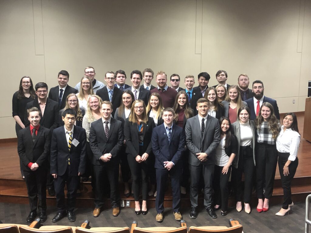 A group of men and women, dressed in formal attire, stand posed in several rows in an indoor setting, likely a lecture hall or conference room.