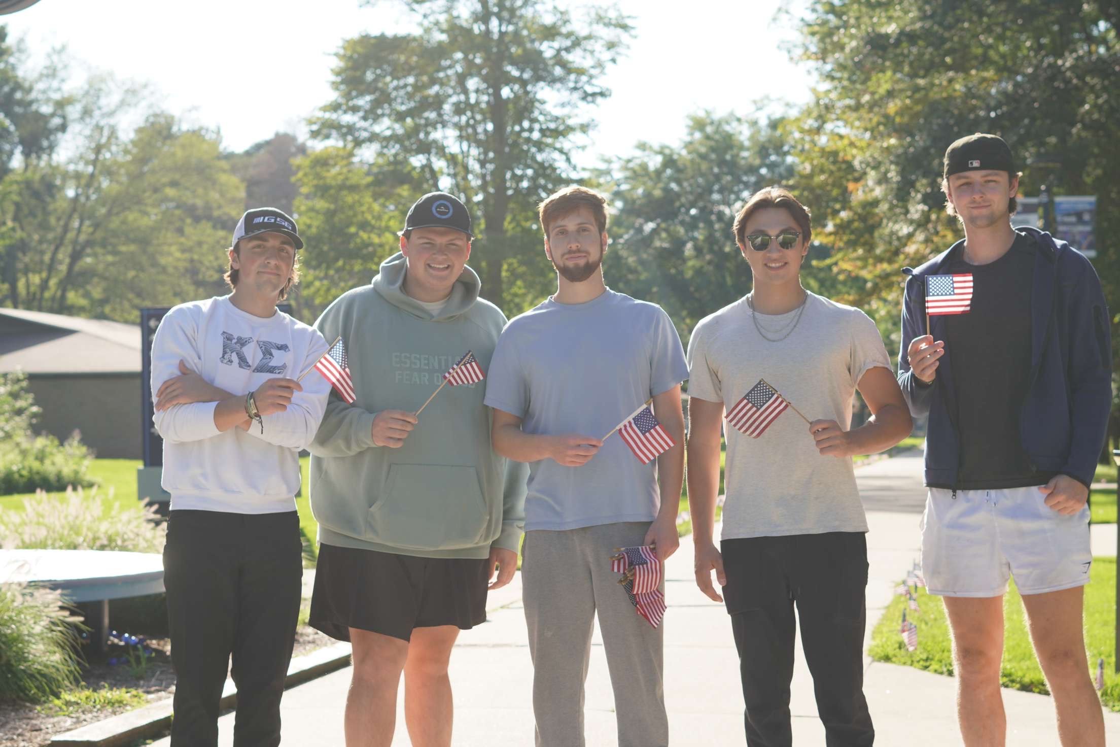 On a sunny day, five men stand outside wearing casual clothing and holding small American flags, honoring the 9/11 anniversary. The scene is a somber yet proud reflection of remembrance, embodying the spirit of unity that Kappa Sigma embraces.
