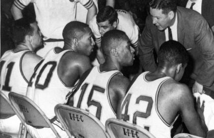 Basketball players in uniform sit on a bench with "K.F.C.C" chairs as a coach and team members discuss strategy during a game. Two referees and a crowd are visible in the background.