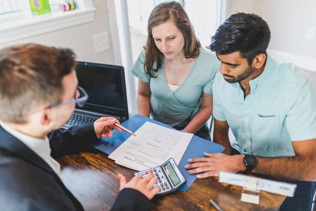 Three people are seated around a table with a laptop, discussing sales management documents. One person is using a calculator.