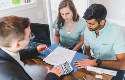 Three people are seated around a table with a laptop, discussing sales management documents. One person is using a calculator.