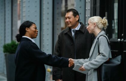 Three people in business attire stand on a city street, two women shaking hands while a man looks on.