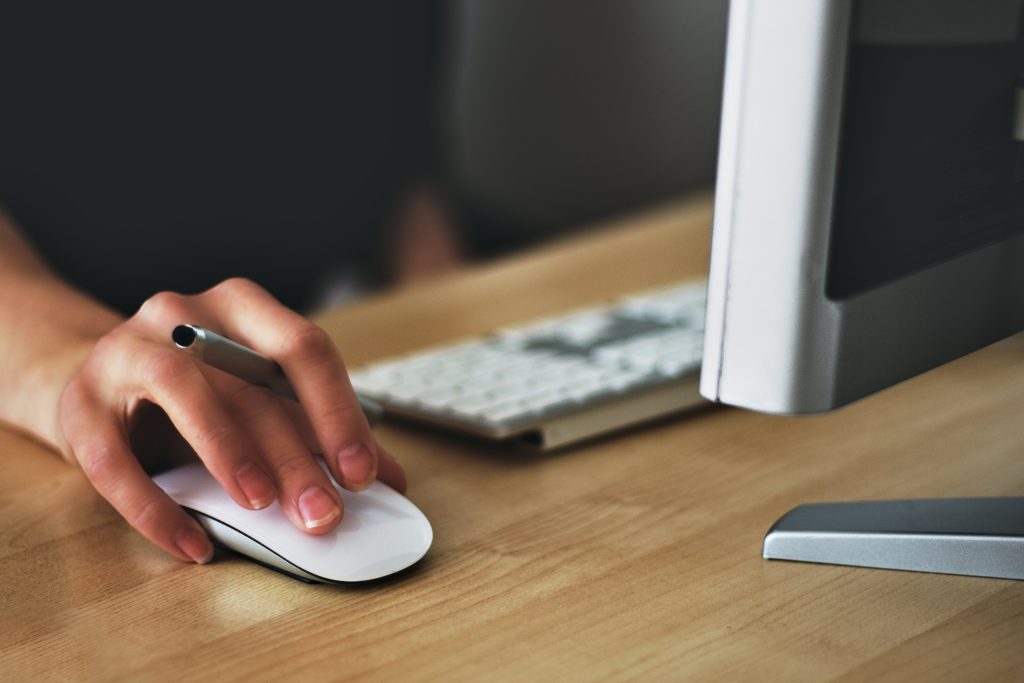 A person's hand navigates a computer mouse on a wooden desk, poised in front of a monitor and keyboard, seamlessly ready for online courses and registration.
