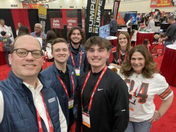 A group of people wearing name badges smiles for a selfie at a convention or trade show with various booths and banners in the background.