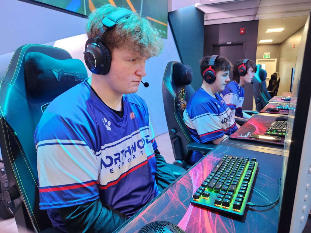 Three individuals are seated at gaming stations, focused on their computer screens, wearing headphones and matching blue jerseys labeled "Northway Esports." Keyboards and gaming mice are visible.