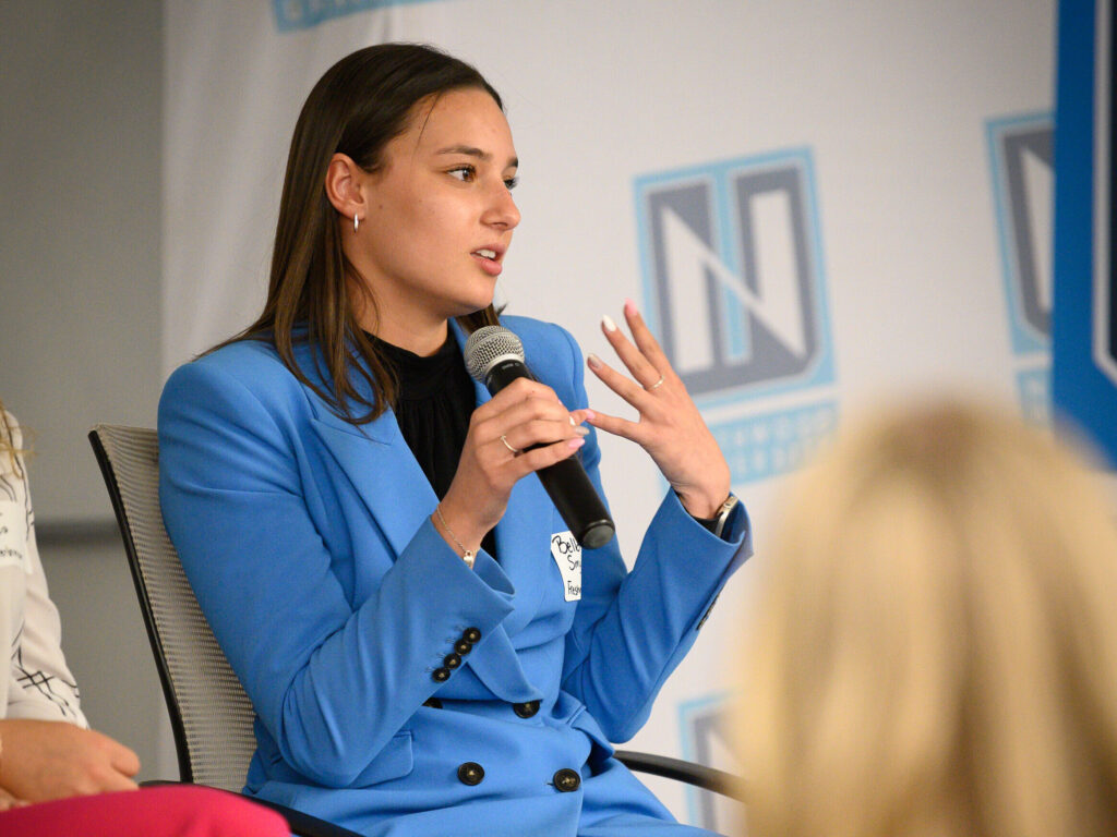 A person wearing a blue blazer speaks into a microphone while gesturing with their other hand during a panel discussion.