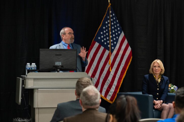 A man speaks at a podium with an American flag behind him, while a woman sits to the side. An audience is seated in the foreground.
