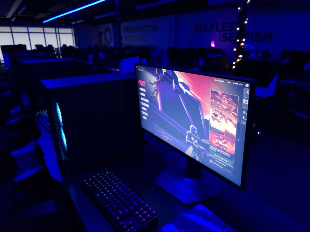 A dimly lit gaming room features a desktop computer with a glowing RGB keyboard and a large monitor displaying a game menu, bathed in blue ambient light.