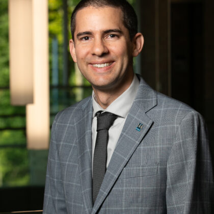 A man wearing a grey suit with a black tie and a pin stands indoors, smiling at the camera. A window with greenery outside is visible in the background.