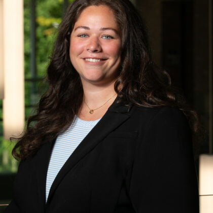 A woman with long brown hair and wearing a black blazer stands indoors, smiling at the camera.