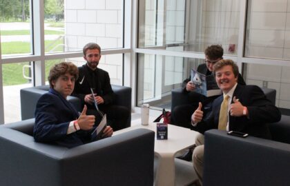 Four men are seated around a white table inside a modern room with glass walls. Three men are giving thumbs up, while one man reads a booklet. They appear to be in a casual or waiting area.