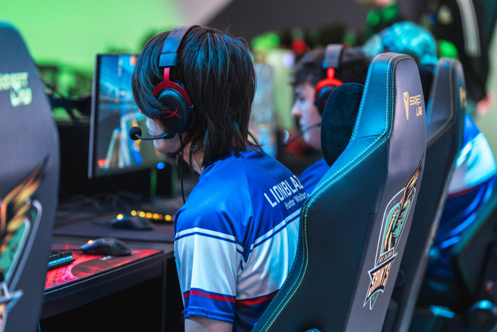 A person wearing a headset and blue jersey sits at a computer desk focused on a monitor in a gaming setup. Two other individuals, also in similar attire, are visible in the background.