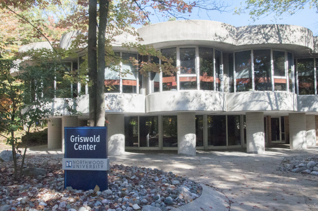 The image shows the Griswold Center at Northwood University. The building has a rounded, modern design with large windows and is surrounded by trees and a landscaped area.