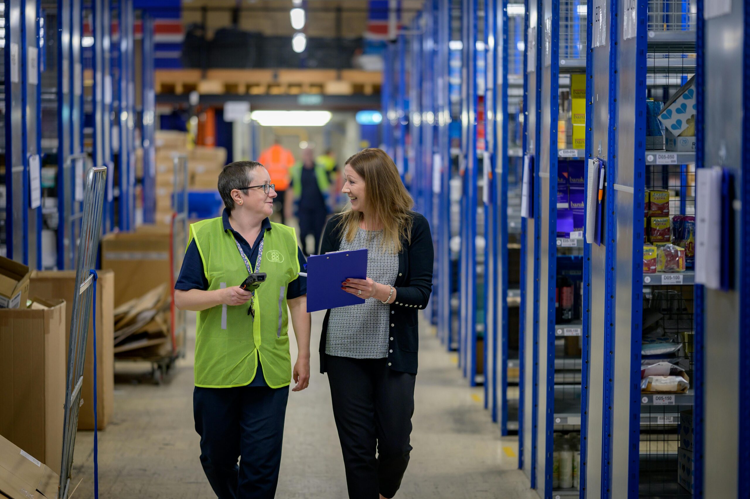 Two women looking at each other and smiling while walking through a warehouse holding clipboards.