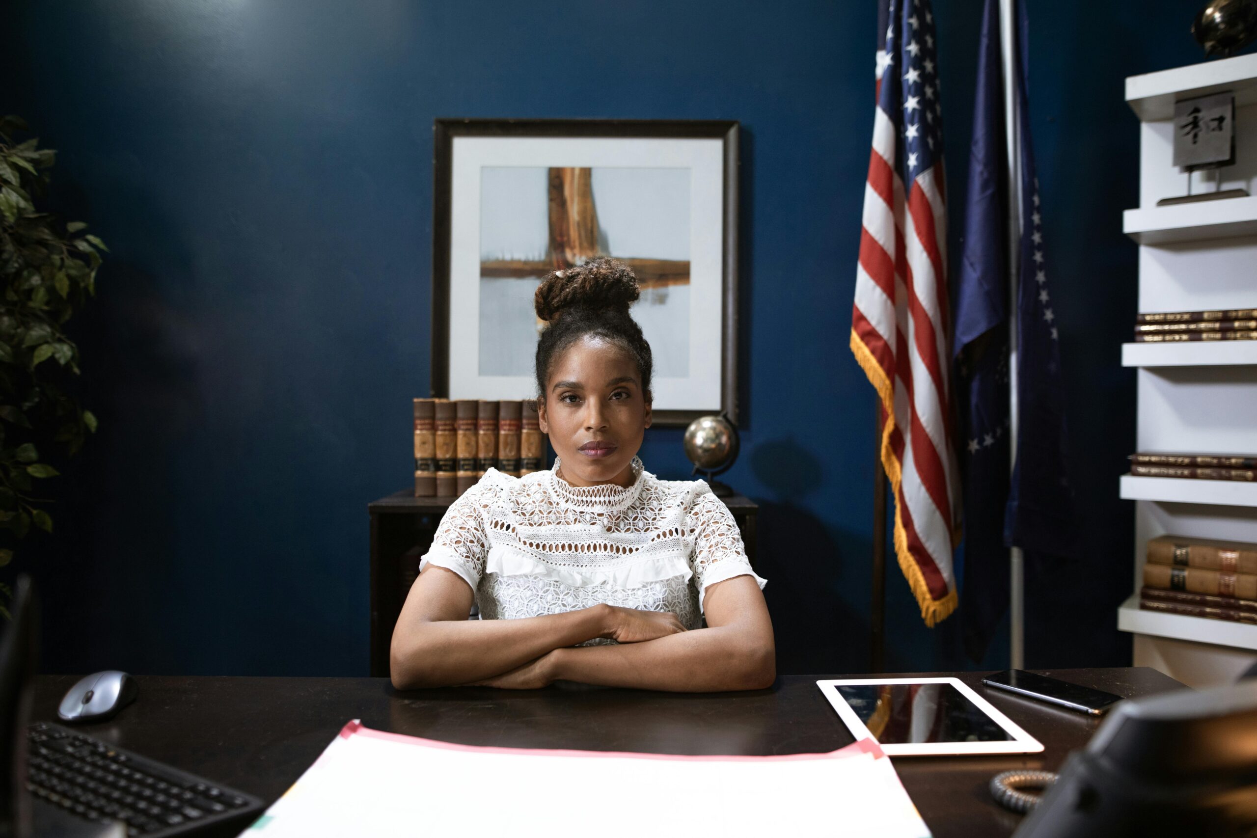 Woman sitting at a wooden desk with papers in front of her, crossing her arms with the american flag in the background.
