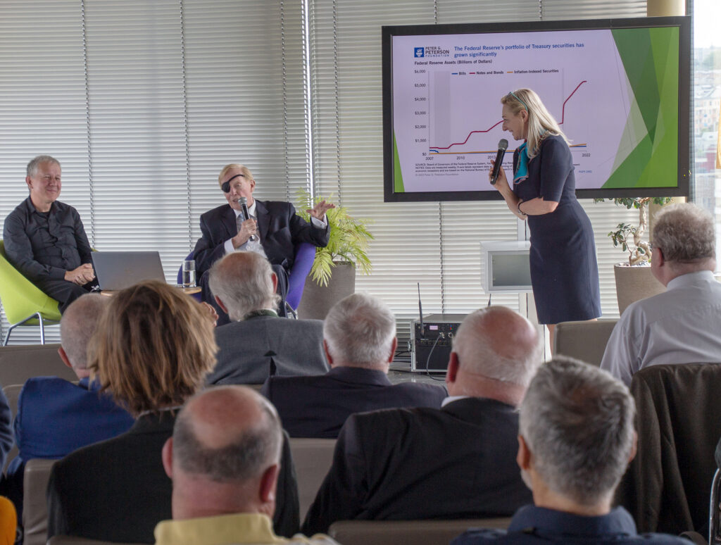 A woman with a microphone speaks to an audience. Behind her, a man gestures towards a screen displaying a graph while seated with another man. The audience is seated and listening attentively.