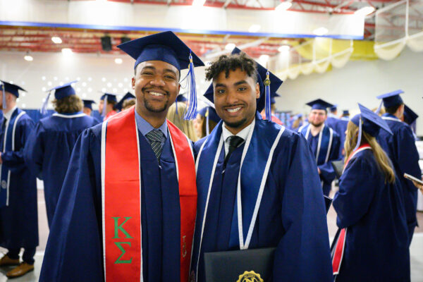 Two men in blue graduation gowns and caps, one wearing a red stole with Greek letters, smile at the camera at a graduation ceremony.