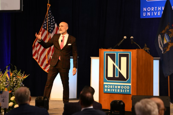 A man in a suit speaks at a podium with a Northwood University logo. An American flag is in the background along with a Michigan state flag. Audience members are seated and watching attentively.