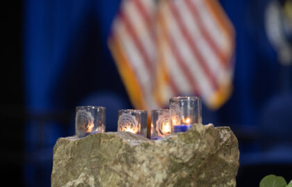 Four lit candles in glass holders rest on a rock with a blurred American flag in the background.