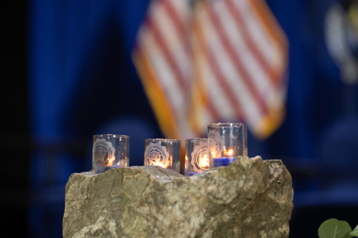 Four lit candles in glass holders rest on a rock with a blurred American flag in the background.
