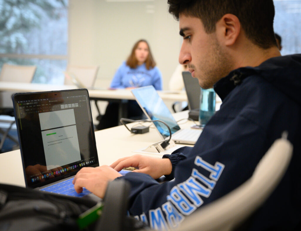 A young man in a blue jacket works on a laptop in a classroom. Other students are seated at desks in the background.