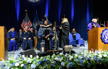 A graduate in academic regalia is hooded on stage during a commencement ceremony, surrounded by university officials and faculty members. American flags and flower decorations are visible in the background.