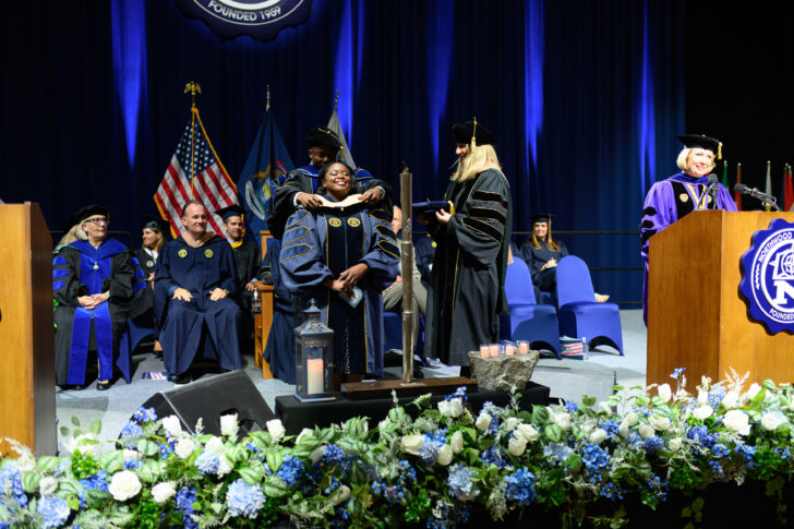 A graduate in academic regalia is hooded on stage during a commencement ceremony, surrounded by university officials and faculty members. American flags and flower decorations are visible in the background.